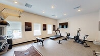 Located at 12556 US-301 in Oxford, Florida, this dedicated exercise room delivers a comprehensive fitness facility within a compact, efficiently utilized single-level footprint. The space features a clean, modern design with neutral white walls, recessed ceiling lighting, and durable carpet flooring suited for high-traffic workout use. A prominent floor-to-ceiling mirror wall visually expands the room while allowing users to monitor their form during exercise. The room is equipped with a RAX multi-functional rack system paired with a tiered dumbbell rack, kettlebell storage, and medicine balls, providing ample options for strength training. Cardio equipment includes a treadmill, a rowing machine, and an air bike, while adjustable weight benches support varied training routines. A wall-mounted flat-screen television offers entertainment during workouts. Natural light enters through several windows along one wall, contributing to an energizing atmosphere. Additional amenities include a wall clock, motivational signage, a water fountain, and a dedicated exit door. The space is well-ventilated with visible ceiling air vents, ensuring comfort throughout the workout experience.