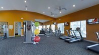 This fitness center is designed to support a full range of workout routines in a bright and energizing environment. The space is defined by bold warm-toned accent walls that create an motivating atmosphere, complemented by neutral carpeted flooring that adds comfort underfoot. Recessed lighting and ceiling fans are installed throughout, ensuring the space remains well-lit and well-ventilated during exercise sessions. Large windows along the walls allow natural light to filter in, contributing to an open and airy feel. The cardio section is equipped with multiple treadmills and elliptical machines, each positioned for easy access and spaced generously to allow comfortable movement. Wall-mounted flat-screen TVs are positioned at viewing height for cardio users, providing entertainment during workouts. A dedicated strength and functional training area features a multi-station cable machine alongside a rack of stability balls in varying sizes. A large floor-to-ceiling mirror spans one wall, enhancing the sense of space while allowing users to monitor their form. The overall layout is thoughtfully organized, offering clearly defined zones for both cardio and strength training within a single, cohesive space.