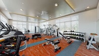 This modern fitness center is designed to support a full range of workout routines in a bright, energizing environment. The space is laid out across a single floor with a distinctive two-toned carpet in bold orange and teal, adding a dynamic visual element throughout. Large floor-to-ceiling windows with horizontal blinds line the walls, flooding the facility with abundant natural light and offering views of an outdoor pool and surrounding greenery.

The main exercise floor is generously proportioned, housing an extensive selection of strength-training equipment, including multiple cable and weight machines, adjustable benches, and a full rack of dumbbells spanning a wide weight range. Functional training is also supported, with equipment such as a tire for battle conditioning exercises. Recessed ceiling lighting and ceiling fans ensure consistent comfort and visibility throughout the space.

A dedicated cardio zone features a row of Matrix treadmills positioned to take advantage of the outdoor views. A separate cycling studio offers spin bikes, wall-mounted mirrors that expand the sense of space, a large flat-screen television, and additional natural light from exterior windows — creating an immersive environment ideal for group or solo cycling sessions. Full-length mirrors throughout the main floor further enhance the open, airy atmosphere of this well-appointed fitness facility.