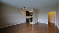 This single-floor space offers a clean, open layout with warm hardwood flooring that extends throughout the main living area, adding a sense of richness and continuity to the interior. The living area is generously proportioned, with neutral-toned walls that provide a versatile backdrop for a variety of interior styles. A track lighting fixture overhead offers directed illumination, enhancing the overall brightness of the space.

Adjacent to the main living area, the kitchen is accessed through an open passage, creating a connected yet defined cooking space. Dark espresso-toned cabinetry lines the kitchen walls, offering ample storage, and includes a distinctive wine rack detail that adds character. The cabinetry is complemented by light countertops, creating a balanced contrast. A stainless steel dishwasher is integrated into the lower cabinetry, contributing to the kitchen's modern functionality. The kitchen floor transitions to a lighter tile surface, further distinguishing it from the living area.

Overall, the space presents a practical and stylish interior that balances open living with a functional kitchen, unified by consistent finishes and a neutral color palette.