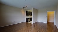 This single-floor space offers a clean, open layout with warm hardwood flooring that extends throughout the main living area, adding a sense of richness and continuity to the interior. The living area is generously proportioned, with neutral-toned walls that provide a versatile backdrop for a variety of interior styles. A track lighting fixture overhead offers directed illumination, enhancing the overall brightness of the space.

Adjacent to the main living area, the kitchen is accessed through an open passage, creating a connected yet defined cooking space. Dark espresso-toned cabinetry lines the kitchen walls, offering ample storage, and includes a distinctive wine rack detail that adds character. The cabinetry is complemented by light countertops, creating a balanced contrast. A stainless steel dishwasher is integrated into the lower cabinetry, contributing to the kitchen's modern functionality. The kitchen floor transitions to a lighter tile surface, further distinguishing it from the living area.

Overall, the space presents a practical and stylish interior that balances open living with a functional kitchen, unified by consistent finishes and a neutral color palette.