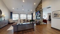 Located at 406 Noel Ln in Smyrna, Tennessee, this single-floor community space delivers a refined yet welcoming atmosphere through a thoughtful blend of rustic and contemporary design elements. The most commanding architectural feature is a floor-to-ceiling stacked-stone fireplace that anchors the main gathering area, fitted with a sleek linear gas insert and a wall-mounted television above — creating a natural focal point for the room. Soaring vaulted ceilings amplify the sense of openness, while rich hardwood flooring flows continuously throughout, grounding the space with warmth and cohesion. The main lounge area is furnished with tufted leather sofas, accent chairs, and a mix of natural wood and metal coffee tables, offering comfortable and versatile seating arrangements. Large windows dressed with patterned curtains and plantation shutters invite abundant natural light while framing views of the surrounding community. A casual dining area features a glass-top high table with dark chairs, positioned near the fireplace and complemented by locally themed black-and-white artwork referencing Smyrna's heritage. Additional seating nooks with tufted accent chairs provide quieter retreats within the space. Recessed lighting throughout ensures a well-lit interior at all hours, and the overall design conveys a sophisticated, community-centered ambiance ideal for gathering and relaxation.