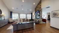 Located at 406 Noel Ln in Smyrna, Tennessee, this single-floor community space delivers a refined yet welcoming atmosphere through a thoughtful blend of rustic and contemporary design elements. The most commanding architectural feature is a floor-to-ceiling stacked-stone fireplace that anchors the main gathering area, fitted with a sleek linear gas insert and a wall-mounted television above — creating a natural focal point for the room. Soaring vaulted ceilings amplify the sense of openness, while rich hardwood flooring flows continuously throughout, grounding the space with warmth and cohesion. The main lounge area is furnished with tufted leather sofas, accent chairs, and a mix of natural wood and metal coffee tables, offering comfortable and versatile seating arrangements. Large windows dressed with patterned curtains and plantation shutters invite abundant natural light while framing views of the surrounding community. A casual dining area features a glass-top high table with dark chairs, positioned near the fireplace and complemented by locally themed black-and-white artwork referencing Smyrna's heritage. Additional seating nooks with tufted accent chairs provide quieter retreats within the space. Recessed lighting throughout ensures a well-lit interior at all hours, and the overall design conveys a sophisticated, community-centered ambiance ideal for gathering and relaxation.