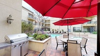 This inviting outdoor courtyard space is thoughtfully designed for both relaxation and socializing. At its center sits a generously sized, rectangular swimming pool with crystal-clear turquoise water, complemented by a separate spa nestled nearby. The pool deck is finished in warm terracotta-toned tiles, giving the area a classic Mediterranean character that carries throughout the space.

Surrounding the pool, a row of lounge chairs provides ample space for sunbathing, while additional seating areas are shaded by bold red patio umbrellas. A built-in BBQ grill station and adjacent dining table with chairs make this courtyard well-suited for outdoor gatherings and casual meals.

Raised planters filled with lush, colorful vegetation — including flowering plants and low-growing succulents — add texture and life to the space. A pergola with decorative columns frames one corner of the courtyard, enhancing its architectural character and offering a shaded retreat.

The entire courtyard is enclosed by wrought iron fencing and bordered by a multi-story building facade featuring balconies above, creating a sense of enclosure and privacy. Mature trees and seasonal blooms throughout the landscaping contribute to the overall ambiance, making this a serene and functional outdoor amenity space.