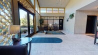 This single-floor space presents a grand and inviting lobby that immediately impresses with its soaring vaulted ceilings and floor-to-ceiling decorative lattice windows, which flood the interior with abundant natural light and offer views of the lush greenery outside. The flooring throughout is a classic marble-style tile that adds an air of elegance and continuity across the space.

The main lobby is thoughtfully arranged into distinct seating areas. One section features a dark upholstered sofa accented with colorful throw pillows, flanked by side tables, positioned directly in front of the striking lattice windows. Nearby, a pair of armchairs with a lamp and side table creates a more intimate conversation nook. A large aquarium serves as a focal point along one wall, complemented by lush indoor trees and framed artwork, adding warmth and life to the environment. Track lighting and a statement chandelier provide a well-lit ambiance throughout.

A bookshelf stocked with reading materials adds a welcoming, community-oriented touch near the corridor entrance. The corridor itself extends deep into the building, lined with overhead flush-mount lighting, textured brick-style wall accents, and dark entry doors, providing clear and functional access to interior spaces. Overall, the space conveys a professional yet comfortable atmosphere suitable for a communal or hospitality setting.