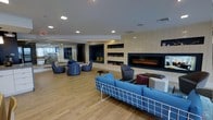 The Library Lounge is a thoughtfully designed social space that seamlessly blends comfort and contemporary style. The open-plan layout is anchored by a striking wide electric fireplace set into a light-paneled feature wall, creating a warm and inviting focal point for the room. Flanking the fireplace are deep-set built-in shelving niches finished in a rich navy blue, offering display space for books and decorative accents that add both color and character to the interior.

The flooring throughout features warm-toned wood planks that lend a grounded, natural feel to the space. Seating is arranged in conversational groupings, including a bold blue grid-frame sofa, patterned blue accent chairs, and a sleek gray lounge chair paired with varied side tables — offering flexibility for both casual gatherings and individual relaxation.

Recessed ceiling lighting provides even, ambient illumination across the space, complemented by a clean white ceiling that enhances the sense of openness. A large mirror along one wall reflects light and visually expands the room. The overall ambiance is modern and welcoming, making the Library Lounge an ideal destination for unwinding, socializing, or browsing a good book in a refined, comfortable environment.