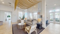 Located in Miramar, Florida, this single-floor space presents a thoughtfully curated coastal aesthetic that blends artistic design with functional layout. The interior is immediately distinguished by its soaring ceilings, which lend an open, airy atmosphere throughout. A dramatic focal point in the main lounge area features an intricate installation of cascading golden rope that drapes from ceiling to floor, creating a striking visual partition while maintaining an open sense of flow between zones.

The lounge area is anchored by soft, neutral furnishings—including plush sofas and accent chairs in light blues and grays—set atop textured area rugs over travertine tile flooring that runs consistently throughout the space. Large floor-to-ceiling windows and glass doors flood every corner with natural light, while offering direct views of an outdoor pool area and surrounding tropical landscaping.

A separate dining area accommodates generous seating beneath oversized woven pendant lights in deep navy, complementing the coastal palette. Curved window bays frame panoramic outdoor views, enhancing the sense of connection to the exterior environment.

The kitchenette is efficiently designed with light wood cabinetry, open shelving, a hexagonal tile backsplash, gray countertops, and stainless steel appliances. The overall ambiance is bright, modern, and resort-inspired throughout.