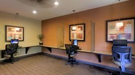 This single-floor space is designed as a dedicated business center, offering a functional and comfortable environment for productivity. The room features a continuous built-in wall-mounted desk that spans the length of the accent wall, providing ample workspace for computing and administrative tasks. A task chair positioned at the central workstation faces a desktop computer, making it a practical hub for focused work.

The warm terracotta-toned accent wall serves as the visual centerpiece of the room, complemented by a framed piece of wall art that adds a polished, professional aesthetic. Glass privacy panels flank the workstation area, creating a sense of semi-private workspaces along the shared desk surface. A pendant light hangs above the central workstation, offering direct illumination, while recessed ceiling lights provide even ambient lighting throughout the space.

The flooring consists of patterned commercial-grade carpet in neutral earth tones, contributing to a quiet and professional atmosphere. A decorative plant placed at the end of the desk adds a touch of warmth and life to the otherwise streamlined workspace. Overall, the space balances utility and comfort, making it well-suited for guests or occupants needing a reliable and well-equipped area to handle business needs.