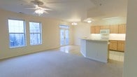 This two-floor space offers an open and airy layout, highlighted by an abundance of natural light streaming through multiple large windows and a set of elegant French doors. The living area features plush carpeting and a ceiling fan, creating a comfortable and functional environment. A chandelier adds a refined touch to the dining area, which transitions seamlessly into the kitchen. The kitchen is defined by a curved breakfast bar peninsula, light wood cabinetry, and white appliances, including a built-in microwave, contributing to a clean and cohesive aesthetic. The flooring in the kitchen and dining area shifts to a smooth hard surface, offering a practical contrast to the carpeted living space. Neutral, warm tones dominate throughout, from the cream-colored walls to the light countertops, lending the space a bright and welcoming atmosphere. The French doors provide access to an outdoor area while also enhancing the flow of natural light into the interior. Overall, the open-concept design and thoughtful layout make this a versatile and well-appointed multi-floor space.