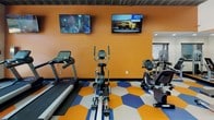 Located at 2424 N Tustin Ave in Santa Ana, California, this single-level fitness center is designed to motivate and energize with its bold, contemporary aesthetic. The space is immediately distinguished by its eye-catching hexagonal floor tiles in vivid shades of orange, blue, and white — a dynamic design element that sets an upbeat, high-energy tone throughout.

The gym is well-equipped with a comprehensive selection of cardio and strength training equipment, including treadmills, elliptical machines, a rowing machine, cable machines, and a full dumbbell rack, accommodating a wide range of fitness routines. Wall-mounted flat-screen TVs are positioned throughout to provide entertainment during workouts.

Exposed ceiling ductwork and linear LED lighting lend an industrial-modern character to the space, while bold blue and orange accent walls reinforce the energetic color palette. An inspirational wall quote — "Follow Your Dreams, Chase The Horizon" — serves as a focal point, adding motivational character to the environment.

Large glass doors and floor-to-ceiling windows along one wall flood the interior with natural light and provide direct views and access to an outdoor area, creating a seamless indoor-outdoor connection. Full-length mirrors are incorporated to support form and technique. The overall ambiance is vibrant, functional, and well-curated for an engaging fitness experience.