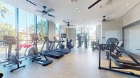 Located at 1611 NW 19th St in Miami, FL, this modern fitness center is thoughtfully designed to provide a complete workout experience in a bright, welcoming environment. The facility occupies a single floor and is characterized by its contemporary architecture, featuring expansive floor-to-ceiling windows that flood the space with natural light and offer views of the surrounding tropical landscaping, including palm trees and lush greenery.

The main workout area is equipped with a full lineup of cardio machines, including multiple treadmills, ellipticals, a rowing machine, and stationary bikes. Ceiling fans are installed throughout, complementing the climate control and ensuring a comfortable workout environment. Light wood-style flooring runs across the entire space, contributing to the clean, modern aesthetic.

A dedicated strength training zone houses a variety of cable and resistance machines designed for full-body conditioning. Full-length mirrors enhance this area, allowing users to monitor their form during exercise. A separate functional training section offers an organized collection of medicine balls, stability balls, resistance bands, foam rollers, and a rack of dumbbells in various weights, catering to dynamic and flexibility-focused workouts. Wall-mounted flat-screen televisions are positioned throughout for entertainment. A water fountain station is also available for convenience.
