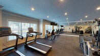 This fitness center is a thoughtfully designed, single-level space that provides a comprehensive workout environment for users of varying fitness levels. The room features recessed lighting paired with natural light from multiple large, white-trimmed windows fitted with blinds, creating a bright and energizing atmosphere. Crown molding details and neutral-toned walls contribute a polished, well-maintained aesthetic throughout.

The cardio section is well-stocked, featuring multiple treadmills, upright and recumbent stationary bikes, ellipticals, and stair climbers arranged to allow comfortable use of each machine. Wall-mounted flat-screen televisions are positioned for viewing from the cardio equipment, enhancing the workout experience. A dedicated strength training zone houses a multi-station cable and weight machine, an adjustable weight bench, and a full rack of dumbbells spanning a wide range of weights. Exercise balls and additional accessories are also available. Floor-to-ceiling mirrors line one wall, amplifying the sense of space and supporting proper form during workouts. Durable rubber flooring covers the entire area, providing safety and noise reduction. Framed artwork and a posted workout schedule add functional and decorative touches to the walls.