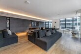 This elegantly designed single-floor lounge space offers a generous, open-plan layout that seamlessly blends comfort with contemporary sophistication. Light oak hardwood flooring flows throughout, complemented by smooth white ceilings with recessed and cove lighting that casts a warm, ambient glow across the entire space.

Floor-to-ceiling windows line the perimeter, flooding the interior with abundant natural light and framing expansive urban views of the surrounding cityscape. The main lounge area features deep, navy-blue and charcoal sectional sofas arranged around low-profile coffee tables, creating multiple inviting conversation zones.

A standout architectural feature is the full-width dark slate feature wall, which houses a sleek, linear gas fireplace — providing both visual drama and warmth. Adjacent to the lounge is a dedicated dining area with a round table and modern chairs, partially separated by striking black-framed frosted glass partition walls that lend an industrial-chic character while maintaining an open feel.

Toward the rear, a well-appointed kitchen bar area is defined by pendant lighting, a dark island with bar seating, and dark cabinetry, offering a refined space for casual dining or entertaining. The overall ambiance is polished, calm, and cosmopolitan — ideal for a premium shared amenity or hospitality environment.