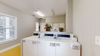 This communal laundry facility offers a clean, efficient, and well-organized space designed to accommodate shared laundry needs. The room is outfitted with multiple coin-operated top-loading washing machines and stacked dryer units, centrally arranged on a raised white platform that keeps machines accessible from multiple sides. The flooring throughout is durable yellow tile, lending the space a bright, utilitarian character. Overhead fluorescent lighting ensures the entire room is well-lit, complementing the natural light that filters in through several windows fitted with blinds, offering views of the surrounding greenery. The walls are painted in a neutral gray tone, creating a clean and orderly atmosphere. A wooden bench near the exit door provides a convenient spot to wait or sort laundry. Safety is addressed with a clearly marked exit door, a fire extinguisher mounted on the wall, and a fire pull station. Decorative touches such as potted floral arrangements add a welcoming feel to the otherwise functional space. The layout provides ample room to move comfortably between machines, making the laundry experience straightforward and efficient for all users.