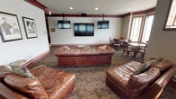 This elegantly appointed amenity suite encompasses three distinct areas — a theater lounge, a billiards room, and a library — all connected by a carpeted corridor with classic architectural detailing including rich wood trim and crown molding throughout. The theater lounge is anchored by a large wall-mounted screen flanked by two additional displays, providing an immersive viewing experience. Plush, tufted leather Chesterfield sofas rest atop a decorative patterned carpet, while framed artwork lines the walls and a bay-windowed dining nook offers additional seating. The billiards room features a full-size pool table with a warm pendant light overhead, decorative wallcovering with a nature-inspired border, and bar-height seating along the wall. The library exudes warmth and charm with striped wallpaper, wood wainscoting, and a fireplace topped by an ornate mantel and a classic portrait painting. Built-in bookshelves, plush seating, and rich drapery complete the inviting atmosphere. A compact kitchenette with dark cabinetry, a refrigerator, microwave, and coffee maker provides convenient refreshment access. Together, these spaces offer a refined and comfortable retreat for leisure and social gathering.