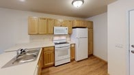 This single-floor residential unit offers a practical and well-organized layout designed for comfortable everyday living. The open-concept living area flows directly into the kitchen, which is separated by a peninsula counter — ideal for casual dining or meal prep. The kitchen is fully equipped with warm honey-toned wood cabinetry providing ample storage, white appliances including a range, over-the-range microwave, and refrigerator, as well as a sink integrated into the countertop. Hard-surface wood-look flooring runs throughout the main living areas and kitchen, offering both durability and a clean aesthetic. The unit includes two bedrooms: one finished with plush carpeting, a ceiling fan with light fixture, and two windows that invite natural light, and a second bedroom with hard-surface flooring, a wall-mounted HVAC unit, and a window with an outdoor view. The bathroom is thoughtfully designed with a walk-in shower stall, toilet, and a wood-toned vanity with storage drawers, complemented by grab bars for added accessibility. The neutral color palette of cream and white walls throughout creates a bright, open ambiance, while consistent wood-tone trim ties the spaces together cohesively.