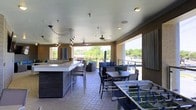 This covered outdoor amenity deck offers a well-appointed social space designed for both relaxation and entertainment. The space is anchored by a sleek bar counter with a light stone surface and dark base, complemented by woven bar stools — ideal for casual gathering or refreshments. A series of stylish pendant lights with an artistic, cascading design hang above the bar area, adding a modern decorative focal point alongside ceiling fans that provide comfortable airflow throughout. The dining area features a contemporary table and chairs set for group meals or casual conversation, while a separate lounge zone offers cushioned seating arrangements with accent pillows for a more relaxed atmosphere. A foosball table adds a recreational element, making the space equally suited for leisure and play. Large open-air sections along the perimeter allow for sweeping views of the surrounding neighborhood and lush tree canopy, flooding the deck with natural light. The tile flooring and neutral-toned walls contribute to a clean, modern aesthetic throughout. Ceiling-mounted speakers and recessed lighting ensure the space is as functional in the evening as it is during the day, creating a versatile environment for a wide range of social activities.