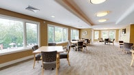 This well-appointed function room offers a welcoming and flexible space designed to accommodate a variety of events and gatherings. The main area is generously sized, featuring multiple seating arrangements with light wood tables and comfortable upholstered chairs set atop neutral, textured carpeting. Warm golden-tan walls and a combination of recessed and flush-mount ceiling lighting create a bright, inviting ambiance throughout.

A defining feature of the room is its expansive bank of large windows along multiple walls, flooding the interior with natural light and providing pleasant views of surrounding greenery and landscaped outdoor areas. The varied ceiling design — including tray ceilings and angled architectural details — adds visual interest and a sense of openness to the space.

A secondary area with a wall-mounted flat-screen television and its own seating arrangement provides a more intimate setting within the larger room, suitable for presentations or small group activities. An adjacent kitchenette supports catering and event needs, equipped with light wood cabinetry, dark countertops, a sink, microwave, and tile flooring — practical and efficiently laid out. Together, these elements make this function room a well-rounded, functional, and aesthetically comfortable venue.
