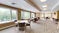This well-appointed function room offers a welcoming and flexible space designed to accommodate a variety of events and gatherings. The main area is generously sized, featuring multiple seating arrangements with light wood tables and comfortable upholstered chairs set atop neutral, textured carpeting. Warm golden-tan walls and a combination of recessed and flush-mount ceiling lighting create a bright, inviting ambiance throughout.

A defining feature of the room is its expansive bank of large windows along multiple walls, flooding the interior with natural light and providing pleasant views of surrounding greenery and landscaped outdoor areas. The varied ceiling design — including tray ceilings and angled architectural details — adds visual interest and a sense of openness to the space.

A secondary area with a wall-mounted flat-screen television and its own seating arrangement provides a more intimate setting within the larger room, suitable for presentations or small group activities. An adjacent kitchenette supports catering and event needs, equipped with light wood cabinetry, dark countertops, a sink, microwave, and tile flooring — practical and efficiently laid out. Together, these elements make this function room a well-rounded, functional, and aesthetically comfortable venue.