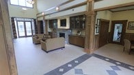 This single-floor space makes a striking impression with its soaring double-height ceilings and open, airy layout. The focal point of the main lounge area is a stacked-stone fireplace flanked by rich, dark-stained built-in cabinetry with glass-front upper display cases, offering both a decorative and functional element to the room. A flat-screen television is mounted above the fireplace, seamlessly integrated into the built-in surround. Sturdy wood beam columns and structural accents lend a warm, craftsman-inspired aesthetic throughout the space. The seating area is furnished with plush, neutral-toned sofas and accent chairs, creating a welcoming and comfortable environment. Underfoot, a refined tile floor features a decorative diamond-pattern border inlay, adding visual interest and a sense of polish to the open floor plan. Natural light filters in through large windows and glass entry doors, enhancing the bright and inviting ambiance. The overall design blends traditional craftsmanship with modern amenities, resulting in a space that feels both functional and elegantly appointed — well-suited for social gatherings or relaxed everyday use.