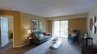 This single-floor residence offers a thoughtfully arranged living space designed for both comfort and functionality. The living room serves as the heart of the home, featuring wall-to-wall carpeting that adds warmth underfoot and a textured ceiling that lends a classic character to the room. Warm, golden-yellow walls create an inviting ambiance, complemented by tasteful wall art that adds a splash of color and personality to the space. A large sliding glass door dominates one wall, flooding the room with abundant natural light and providing direct access to an outdoor balcony. The view through the glass hints at lush greenery beyond, offering a refreshing connection to the outdoors. The living area is well-proportioned, comfortably accommodating a tufted sofa accented with colorful throw pillows, a coordinating accent chair, and a sleek glass coffee table. A side table with a table lamp adds a soft, ambient glow for evening use. The overall design balances casual comfort with clean, simple lines, making the space feel open, airy, and livable. The layout is practical and efficient, with an easy flow suited for everyday living.