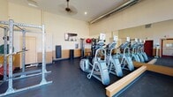 Located at 2160 Rice Lake Rd in Duluth, Minnesota, this single-floor fitness center is designed to support a well-rounded workout experience. The space features soaring ceilings with exposed ductwork that lend it an open, industrial character, while warm neutral tones on the walls and two-tone wainscoting create an inviting atmosphere.

The cardio section is well-equipped with multiple treadmills, elliptical trainers, and stair climbers from Precor, all arranged along a wall of floor-to-ceiling mirrors that amplify the sense of space. Large, multi-pane windows along one wall flood the area with natural light and offer views of the surrounding community. A ceiling fan overhead aids in air circulation during workouts.

The strength training area includes a heavy-duty power rack loaded with a full set of color-coded weight plates, as well as a functional cable machine and a dedicated free weights section with a dumbbell rack. A flat-screen television is mounted on the wall for entertainment. Durable rubber flooring covers the workout areas, providing safety and support. A body weight scale near the entrance offers a convenient health monitoring station. The fitness center is accessible via a clearly marked exit door and is well-lit throughout, making it a functional and comfortable facility for daily exercise.