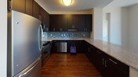 Located at 11215 Georgia Ave in Silver Spring, Maryland, this well-appointed high-rise unit offers a thoughtfully designed single-floor layout with a mix of stylish finishes and functional spaces. The open-concept living and dining area features rich, dark hardwood flooring and large floor-to-ceiling windows that flood the space with natural light while providing sweeping panoramic views of the Silver Spring skyline and surrounding landscape. A built-in media niche adds a distinctive architectural detail to the main living area. The kitchen is outfitted with dark espresso cabinetry, granite countertops, stainless steel appliances including a refrigerator, range, and dishwasher, and a decorative mosaic tile backsplash that adds a contemporary touch. The bedrooms are finished with plush wall-to-wall carpeting and feature large windows that continue the theme of abundant natural light and elevated city views. The bathrooms are clean and modern, featuring tile flooring, soaking tubs with tiled surrounds, granite-topped dark vanities, and chrome fixtures. The neutral color palette throughout the unit creates a bright, cohesive ambiance that complements both the interior finishes and the expansive urban views beyond.