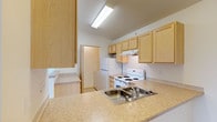 This single-floor space is designed with practicality and comfort in mind, offering a clean and functional layout throughout. The kitchen serves as a focal point of the home, showcasing light natural wood cabinetry that lines the walls and provides generous storage space. The countertops offer ample room for food preparation, complemented by a double-basin stainless steel sink with a convenient faucet. Essential appliances are included, featuring a white electric range with a ventilation hood above, a full-size refrigerator, and an additional stacked appliance unit, likely a washer and dryer, tucked neatly into a dedicated alcove. Overhead fluorescent lighting ensures the kitchen remains bright and well-lit. A pass-through counter connects the kitchen to an adjacent area, enhancing the sense of open flow within the space. The neutral-toned walls and light finishes throughout the kitchen create a clean, airy atmosphere. A doorway visible from the kitchen leads further into the unit, hinting at additional living areas beyond. The overall design prioritizes efficient use of space, making it a practical and inviting environment for daily living.