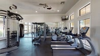 Located at 1200 W Corral Ave in Kingsville, Texas, this fitness center is a single-floor facility designed to support a comprehensive range of workout routines. The space is characterized by elevated ceilings with crown molding detail, recessed lighting, and a ceiling fan that contributes to a comfortable exercise environment. Large, multi-pane windows line the walls, flooding the interior with abundant natural light and offering views of the surrounding outdoor area, including a glimpse of a pool and landscaping beyond.

The flooring throughout is a dark, rubberized material suited for high-impact activity. Cardio equipment is well-represented, with multiple treadmills, elliptical machines, and stationary bikes positioned to take advantage of the natural light from the windows. A large wall-mounted mirror spans one side of the room, visually expanding the space and allowing users to monitor their form.

The strength training section includes a multi-functional cable machine, weight benches, a dumbbell rack with various weighted options, and a power rack with pull-up bar. A hanging punching bag and battle ropes add functional training options to the mix. Stability balls and resistance accessories are stored along one wall. A wall-mounted television provides entertainment, and French doors connect the space to an adjacent area, enhancing accessibility throughout the facility.