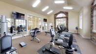 Located in Puyallup, WA, this well-appointed fitness center offers a spacious, single-floor layout designed to support a comprehensive workout experience. The room's vaulted ceiling rises to a dramatic peak, creating an open, airy atmosphere that enhances the sense of space throughout. A standout architectural feature is the stone-accented arched window focal wall, where stacked natural stone surrounds a large arched transom window, flooding the space with natural light and adding a refined, upscale aesthetic.

Large multi-pane windows line additional walls, further brightening the interior and providing views of the surrounding landscape. The warm, neutral-toned walls and carpeted flooring contribute to a comfortable and inviting environment. Wall-mounted flat-screen televisions are positioned throughout for entertainment during workouts.

The fitness center is equipped with a thorough selection of exercise machines, including treadmills, elliptical trainers, stationary bikes, and multi-station strength training equipment, as well as free weights and benches. Full-length mirrors along one wall allow for proper form monitoring and visually expand the room. The layout is thoughtfully organized to provide ample space between equipment, ensuring ease of movement and a safe, functional workout environment for all users.