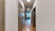 This single-floor residence is thoughtfully designed with a cohesive, modern aesthetic throughout. A central hallway serves as the spine of the unit, lined with taupe-finished panel doors and illuminated by recessed ceiling lights. The hardwood-style flooring runs continuously through the main living and kitchen areas, lending warmth and visual continuity to the space.

The kitchen is well-appointed, featuring dark espresso cabinetry paired with light granite countertops and a subtle tile backsplash. Stainless steel appliances — including a range, over-the-range microwave, refrigerator, and dishwasher — complete the functional and stylish cooking space. A ceiling fan is visible in the adjacent living area, which benefits from a large window offering views of surrounding greenery.

The bedrooms offer a calm, neutral palette with carpet flooring in at least one room and hardwood-style flooring in another. Windows with horizontal blinds allow natural light to filter in while maintaining privacy. A bedroom with three tall windows is particularly bright, providing an inviting sense of openness.

The bathroom includes a wide vanity with white cabinetry, granite countertop, an under-mount sink, and a large framed mirror with overhead lighting. The overall design is clean and consistent, with neutral tones, modern finishes, and practical layouts throughout.