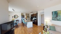 This single-floor residence offers a thoughtfully designed open-concept layout that seamlessly connects the kitchen, dining, and living areas. The kitchen is a standout feature, equipped with dark espresso cabinetry, granite countertops, a center island with bar seating, stainless steel refrigerator, and black appliances illuminated by track lighting overhead. Warm hardwood-style flooring flows throughout the main living spaces, enhancing the bright and airy atmosphere created by abundant natural light from large windows dressed with teal drapes.

The living room is generously sized, with a glass-panel door providing access to an outdoor area and views of the surrounding greenery. Adjacent, the dining area accommodates a full dining set beneath large windows that invite natural light and outdoor views.

The residence includes two bedrooms, each carpeted for comfort and featuring multiple windows with views of the outdoors. One bedroom is notably spacious with a triple-window configuration. Both bathrooms are well-appointed — one features a soaking tub with tile flooring and a stone-topped vanity, while the second offers a full tub-shower combination with dark overhead cabinetry. The overall ambiance is modern, clean, and inviting throughout.
