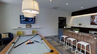 This contemporary single-floor amenity space is thoughtfully designed to serve as a social and recreational hub. The open-plan layout centers around a full-size pool table with a light gray felt surface, positioned beneath a pair of large, modern drum pendant lights that cast a warm, inviting glow throughout the room. A wall-mounted flat-screen TV is positioned above a floating shelf, creating a comfortable entertainment area complemented by nearby seating with sleek, modern chairs. The flooring features a stylish multi-toned geometric carpet tile pattern that adds visual depth and defines the space. Toward the rear, a bar-style counter with a warm wood finish is paired with contemporary white bar stools, offering a casual gathering spot. Adjacent cabinetry with dark accents and under-cabinet lighting adds a refined touch, while a small potted plant introduces a subtle natural element. The walls are finished in clean white, enhancing the bright and airy atmosphere. Recessed lighting throughout supplements the pendant fixtures, ensuring the space is well-lit and welcoming. Overall, the design strikes a balance between modern aesthetics and functional comfort, making it an ideal setting for leisure and community interaction.