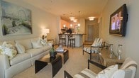 This single-floor residence showcases an open-concept layout that connects the living area and kitchen in a seamless, functional flow. The living space is anchored by a neutral-toned sofa and complementary accent chairs upholstered in a classic stripe pattern, arranged around a sleek dark coffee table. Soft carpeting underfoot adds warmth and comfort to the space.

The kitchen sits at the heart of the home, featuring a peninsula-style counter with bar-height seating for three, offering both a casual dining area and a natural division between spaces. Dark appliances and warm cabinetry create a contemporary contrast against the light-colored walls. A cluster of pendant lights in varying shapes hangs above the kitchen area, while additional pendant and overhead fixtures illuminate the space with a warm, inviting glow throughout.

The entry door is visible in the background, framing the layout clearly. Neutral wall tones and consistent warm lighting contribute to a cohesive, welcoming ambiance across the entire space. Decorative accents, including potted greenery, framed wall art, and layered throw pillows, add personality without overwhelming the clean aesthetic.