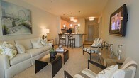 This single-floor residence showcases an open-concept layout that connects the living area and kitchen in a seamless, functional flow. The living space is anchored by a neutral-toned sofa and complementary accent chairs upholstered in a classic stripe pattern, arranged around a sleek dark coffee table. Soft carpeting underfoot adds warmth and comfort to the space.

The kitchen sits at the heart of the home, featuring a peninsula-style counter with bar-height seating for three, offering both a casual dining area and a natural division between spaces. Dark appliances and warm cabinetry create a contemporary contrast against the light-colored walls. A cluster of pendant lights in varying shapes hangs above the kitchen area, while additional pendant and overhead fixtures illuminate the space with a warm, inviting glow throughout.

The entry door is visible in the background, framing the layout clearly. Neutral wall tones and consistent warm lighting contribute to a cohesive, welcoming ambiance across the entire space. Decorative accents, including potted greenery, framed wall art, and layered throw pillows, add personality without overwhelming the clean aesthetic.
