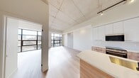 This thoughtfully designed 1-bedroom, 2-bathroom unit delivers a clean, contemporary aesthetic throughout its efficiently organized single floor. The open-concept living and kitchen area is immediately striking, featuring soaring exposed concrete ceilings with industrial-style track lighting that contrast elegantly with crisp white walls and light-toned wide-plank hardwood floors. Large floor-to-ceiling windows along one wall flood the living space with natural light and provide views of the surrounding area.

The kitchen is a standout feature, centered around a generous waterfall-edge island with an undermount sink and matte black faucet. Cabinetry blends white upper units with warm wood-toned lower cabinets, complemented by a textured tile backsplash and stainless steel appliances including a refrigerator, range, and built-in microwave.

The bedroom is a comfortable, well-proportioned room with light wood flooring, a narrow vertical window, and an exposed concrete structural column that adds an industrial character. Three hallways connect the private areas of the unit seamlessly.

The two bathrooms offer distinct configurations: one features a full soaker tub with glossy white tile surround, while the second includes a stand-up glass-enclosed shower, white vanity with under-cabinet storage, and terrazzo-style tile flooring with a warm brass wall sconce. Both are finished in a clean, minimalist style consistent with the overall design language of the space.