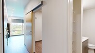 Located at 1411 E Iron Bridge Way in Spokane, Washington, this efficiently designed unit offers a thoughtfully organized single-floor layout. The open-plan kitchen serves as a central feature, outfitted with bold blue upper cabinetry contrasted by light grey lower cabinets and white countertops. Stainless steel appliances — including a refrigerator, range, microwave, and dishwasher — provide a full suite of modern conveniences. Recessed lighting illuminates the kitchen, which is anchored by a large picture window offering expansive views of surrounding greenery, flooding the space with natural light. Light-toned wood-look flooring runs throughout, lending warmth and visual continuity to each area. The living area is open and clean-lined, with neutral white walls and dark baseboards creating a crisp, contemporary aesthetic. The hallway, featuring a sleek sliding door with a gold-toned frame, connects the living areas to the bathroom. The bathroom is well-appointed with a grey vanity, white countertop, mirror, and a stacked washer and dryer unit, combining laundry convenience with a clean, functional design. Overall, the space presents a modern, minimalist character with practical finishes and a bright, airy atmosphere.
