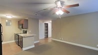 This single-floor space is designed with an open-concept layout that seamlessly connects the kitchen and living area, creating a cohesive and functional environment. The living area features neutral-toned carpet flooring and warm beige walls, contributing to a calm and understated ambiance. A ceiling fan with an integrated light fixture is centrally positioned overhead, providing both air circulation and ambient lighting. Track lighting runs along the ceiling, offering additional illumination options.

The kitchen is situated along one wall and is partially separated from the living space by a peninsula counter with a light-toned surface, which provides a casual dining or prep area. Dark wood cabinetry lines the kitchen walls, complemented by a stainless steel range, creating a functional cooking space with a defined aesthetic.

A notable feature of this unit is the large walk-in closet visible through an open door, equipped with built-in shelving units that offer generous storage capacity. The open doorway creates visual depth in the space. White baseboards and trim throughout provide clean contrast against the neutral walls, lending a polished, finished look to the overall interior.