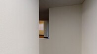 This single-floor unit offers a well-organized layout that balances functionality with comfort. The living area is a generous, open space anchored by a clean, white-surround fireplace with a simple mantel, complemented by rich, dark hardwood-style flooring and vertical blinds that allow for controlled natural light. A ceiling fan adds practicality to the living space, which flows seamlessly toward the kitchen area.

The kitchen features an open, pass-through design with a raised breakfast bar counter finished in dark laminate, providing a natural dividing point between the kitchen and living areas. Light wood cabinetry lines the kitchen walls, paired with black appliances including a refrigerator, range, and microwave. Overhead lighting illuminates the workspace effectively.

The bedroom is a well-proportioned, quiet retreat finished with neutral carpet and a large window fitted with vertical blinds, offering natural light and privacy. A baseboard heater runs along the wall beneath the window. The walls throughout the unit are painted in soft, neutral tones, creating a clean and cohesive aesthetic.

The dollhouse view reveals a compact, efficient floor plan with clearly defined zones for living, cooking, and sleeping, along with what appears to be a bathroom and additional storage areas, making this a practical and comfortable space.