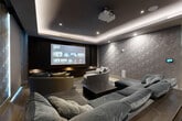 Located in Fort Lauderdale, FL, this dedicated home theater room delivers a refined cinematic experience through thoughtful design and premium finishes. The space is anchored by a large-format projection screen set against a dark, wood-paneled feature wall with integrated illuminated display niches, creating a true theater focal point. A ceiling-mounted projector delivers sharp, immersive visuals, while in-ceiling speakers ensure high-quality audio throughout. The tray ceiling is outfitted with recessed LED strip lighting that casts a warm, ambient glow, enhancing the room's intimate atmosphere. Geometric three-dimensional wall panels in a cool grey tone line the surrounding walls, adding a striking textural element that also aids in acoustic performance. Light hardwood flooring runs throughout and is partially covered by a bold chevron-patterned area rug, grounding the seating arrangement. Generous, deeply cushioned sectional sofas in dark grey velvet are complemented by a rounded, oversized accent chair in lighter grey upholstery, offering ample and comfortable seating for a full audience. Motorized blackout roller shades on the windows allow for complete light control. A smart home control panel is integrated into the wall, enabling seamless management of the room's systems. The overall ambiance is dark, sophisticated, and purpose-built for an exceptional viewing experience.