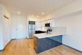Located on 6th Avenue in Tacoma's vibrant North End, this modern one-bedroom, one-bathroom unit offers a well-designed open-concept layout finished with light hardwood flooring throughout. The combined kitchen and living area serves as the heart of the unit, where a sleek peninsula with a textured blue face and white quartz countertop provides both prep space and a casual seating area. The kitchen is outfitted with a full suite of stainless steel appliances — including a refrigerator, electric range, over-the-range microwave, and dishwasher — complemented by two-toned cabinetry featuring dark lower cabinets and crisp white upper cabinets with a tile backsplash. The living area is bright and open, with large sliding glass doors leading to a private balcony that overlooks the surrounding neighborhood, bringing in generous natural light. The bedroom is a comfortable size, featuring a large picture window with neighborhood views, a walk-in closet with built-in shelving, and the same warm hardwood flooring. The bathroom includes a dark wood vanity with a white countertop, a large mirror, and a clean, modern finish. Recessed lighting and a neutral color palette complete the contemporary aesthetic throughout.