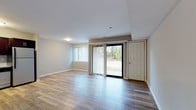 This single-floor unit offers a thoughtfully designed open-concept layout with a clean, modern aesthetic throughout. The spacious living area is finished with light-toned hardwood-style flooring that extends seamlessly across the main space, creating a bright and cohesive feel. Large sliding glass doors are flanked by vertical blinds and a side entry door, allowing natural light to flow in while providing direct access to the outdoors and views of the surrounding greenery.

The kitchen is positioned in an alcove off the main living area and is outfitted with rich dark espresso cabinetry, granite countertops with subtle grey and white veining, and a light grey subway tile backsplash. Stainless steel appliances—including a refrigerator, electric range, built-in microwave, and dishwasher—complete the functional and stylish cooking space.

The bathroom features a clean, neutral palette with large-format white floor tiles, a dark espresso vanity topped with matching granite countertops, and a full tub-shower combination with tiled surround. White panel doors and crisp white trim are consistent throughout the unit, contributing to a polished, uniform finish. The overall ambiance is modern, clean, and move-in ready.