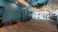 This fitness center is designed to support a full range of workout routines within a single, open floor layout. The space makes an immediate visual impression with its striking color palette — vibrant teal and bold orange accent walls paired with a deep navy tray ceiling — creating an energetic and motivating atmosphere. Recessed lighting and a large ceiling fan ensure the environment remains well-lit and comfortable throughout.

The flooring throughout is a durable, textured carpet tile in earthy tones with blue accents, providing both function and style. A wall of mirrors anchors one end of the room, visually expanding the space while allowing exercisers to monitor their form.

The equipment selection is comprehensive. A dedicated free weights zone features a fully stocked dumbbell rack set against the mirrored wall, accompanied by a power rack and weight bench for strength training. Cardio options include treadmills positioned along a large window wall that brings in natural light, as well as stationary bikes and an elliptical-style machine. A boxing station with a heavy Everlast punching bag, speed bags, and accessory rack rounds out the offering. Additional equipment such as a freestanding multi-use strength machine and a padded mat complete the versatile layout, making this fitness center suitable for a wide range of training styles and fitness levels.
