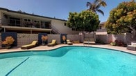 This inviting outdoor amenity space is centered around a generously sized freeform swimming pool with crystal-clear blue water, surrounded by a wide concrete pool deck designed for both relaxation and socializing. The pool area is thoughtfully appointed with cushioned chaise lounges and rattan seating, ideal for sunbathing under the abundant natural light and open sky.

Decorative breeze-block walls frame portions of the perimeter, lending a mid-century modern aesthetic that complements the overall design of the surrounding building. Mature palm trees and lush ornamental plantings, including vibrant potted specimens, add a tropical, resort-like ambiance to the setting.

A covered outdoor patio area is enclosed with full-height glass panels, featuring a warm wood-paneled ceiling, recessed lighting, and a wall-mounted television, creating a shaded lounge space that connects seamlessly to the pool deck. Additional seating arrangements with cushioned sofas and accent chairs complete this entertainment zone.

The building backdrop features an upper-level balcony with staircase access, reinforcing the communal, multi-unit character of the property. The combination of the sparkling pool, stylish breeze-block detailing, lush greenery, and glass-enclosed lounge creates a cohesive and well-appointed outdoor living environment suited for year-round enjoyment in a warm, sunny climate.