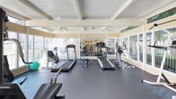 This fitness center is designed to support a comprehensive workout experience within a bright, open single-floor layout. The main exercise area features rubber-matted flooring throughout, providing a durable and slip-resistant surface suitable for a wide range of activities. Large windows line multiple walls, flooding the space with natural light and offering views of the surrounding outdoor greenery and pool area. Ceiling fans are mounted to the white-paneled, beam-accented ceiling, ensuring consistent airflow during workouts.

The cardio zone is equipped with multiple treadmills and an elliptical trainer, complemented by a rowing machine and stationary bike. A full-length wall mirror expands the sense of space and allows users to monitor their form. A dedicated area accommodates strength training with a multi-station cable machine, weight bench, and free weights. A separate section features a standing punching bag and pull-up station on protective matting, ideal for martial arts and functional training.

A small kitchenette area with hardwood-style flooring, a sink, a full-size refrigerator, a compact refrigerator, and a water cooler provides convenient amenities for post-workout refreshment.