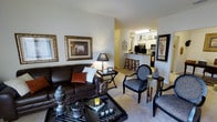 This single-floor residential space offers a thoughtfully designed open-concept layout that seamlessly connects the main living area to the kitchen, creating a sense of flow and accessibility throughout. The living room serves as the heart of the space, featuring plush carpeted flooring and ample room for comfortable seating arrangements. Warm, neutral-toned walls provide a versatile backdrop, complemented by decorative framed artwork that adds character and visual interest. The space is well-lit with a combination of natural light from windows and ambient interior lighting. A stylish coffee table anchors the seating area, which is accented with eclectic furnishings that lend a refined, curated aesthetic. Adjacent to the living area, the kitchen is visible through a convenient pass-through opening, complete with a raised breakfast bar and bar stools — ideal for casual dining or entertaining. The kitchen is equipped with standard appliances, including a refrigerator and microwave, set against dark cabinetry that adds a bold contrast to the lighter surroundings. The overall ambiance of the space is warm and inviting, blending functional design with decorative detail to create a comfortable and stylish single-floor living environment.
