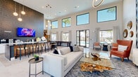 Located at 105 Mazari Ln in Venice, Florida, this single-story community clubhouse offers a sophisticated and welcoming gathering space defined by striking contemporary design. The heart of the facility is a dramatic great room with soaring double-height ceilings and an expansive bank of clerestory windows that flood the interior with natural light, offering views of the surrounding Florida landscape and palm trees. A bold, textured geometric accent wall anchors the main lounge area, which is appointed with stylish seating arrangements including plush sofas, accent chairs, and glass-top coffee tables. A large wall-mounted TV serves as a focal point for entertainment. The central bar area features a sleek dark countertop with modern wishbone bar stools, complemented by a white cabinetry service station and pendant globe lighting above. A shuffleboard table adds a recreational element to the space. A long, light-filled corridor with black-framed glass doors and large windows connects various areas of the facility, including semi-private nook seating and additional lounge rooms adorned with textured 3D wall panels. The light tile flooring, warm wood beam accents, and curated artwork throughout contribute to a refined yet approachable ambiance, making this clubhouse an inviting destination for community gatherings.