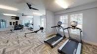 This fitness center offers a thoughtfully designed workout environment spread across a single open floor. The space is finished with neutral-toned patterned carpet tiles that lend a modern aesthetic while providing underfoot comfort throughout the workout area. Soft blue-gray walls and bright flush-mount lighting fixtures create a clean, energizing atmosphere, complemented by ceiling fans that help maintain air circulation during exercise sessions.

Large windows along multiple walls allow abundant natural light to fill the space, offering views of the surrounding outdoors. Full-length mirrors are strategically placed to expand the visual depth of the room and assist users during workouts. The layout is well-organized, with distinct zones for cardio and strength training.

The cardio section includes True-brand treadmills, elliptical machines, a rowing machine, and stationary bikes, while the strength area features a cable multi-use machine, a rack of dumbbells, and an adjustable weight bench. A wall-mounted flat-screen television provides entertainment for users. French doors serve as an additional entry point, enhancing the open, accessible feel of the facility.