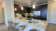 This single-floor space centers around a well-appointed, open-concept kitchen that blends contemporary design with practical functionality. Dark espresso cabinetry lines the walls from base to ceiling, providing generous storage while creating a bold visual contrast against the light granite countertops. The countertops feature warm gold and cream tones with subtle veining, lending an upscale aesthetic throughout.

A wide breakfast bar extends the counter space, offering seating for multiple guests on stylish wooden bar stools with metal bases — ideal for casual dining or entertaining. Above the bar, a series of pendant lights with cylindrical glass shades hang from a track, casting warm, focused light over the surface and enhancing the inviting atmosphere.

The kitchen is fully equipped with stainless steel appliances, including a French door refrigerator, a built-in microwave, and a range, all complemented by a light green subway tile backsplash that adds a subtle pop of color. Under-cabinet lighting further illuminates the workspace. Track lighting along the ceiling ensures the entire area is well-lit, making this kitchen as functional as it is visually appealing.