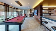 This single-floor amenity lounge is thoughtfully designed to serve as a dynamic social and entertainment hub. The space centers around a full-size pool table with a bold red felt surface, positioned beneath a sleek pendant light fixture that anchors the room's layout. Adjacent to the pool table, a shuffleboard table sits along a warm, earth-toned accent wall adorned with decorative laser-cut panels, framed artwork, and a floating shelf with greenery — all illuminated by stylish industrial pendant lights.

A rich wood-paneled feature wall houses a large flat-screen television alongside bar-height seating, creating a dedicated media and lounge area. The wet bar and kitchen nook behind it offer additional functionality for gatherings. Patterned carpet tiles in a neutral herringbone design flow throughout the space, complementing the warm interior palette.

Floor-to-ceiling windows along one side flood the room with natural light and provide expansive views of the surrounding outdoor green spaces and neighboring buildings. High-top tables with cushioned red chairs and casual seating arrangements throughout the space ensure ample room for socializing. The overall ambiance strikes a balance between modern sophistication and relaxed comfort, making it an inviting destination for both casual hangouts and organized events.